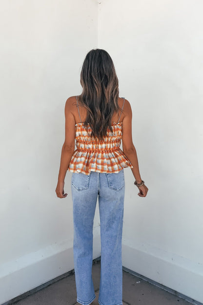 Woman with wavy brown hair wears the Aureum Multi Plaid Smock Top, standing by a white corner wall in light blue jeans.