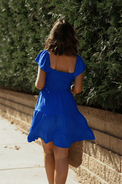 A woman in Ava’s Sweetheart Blue Mini Dress walks by green bushes, holding a woven bag, her back to the camera.