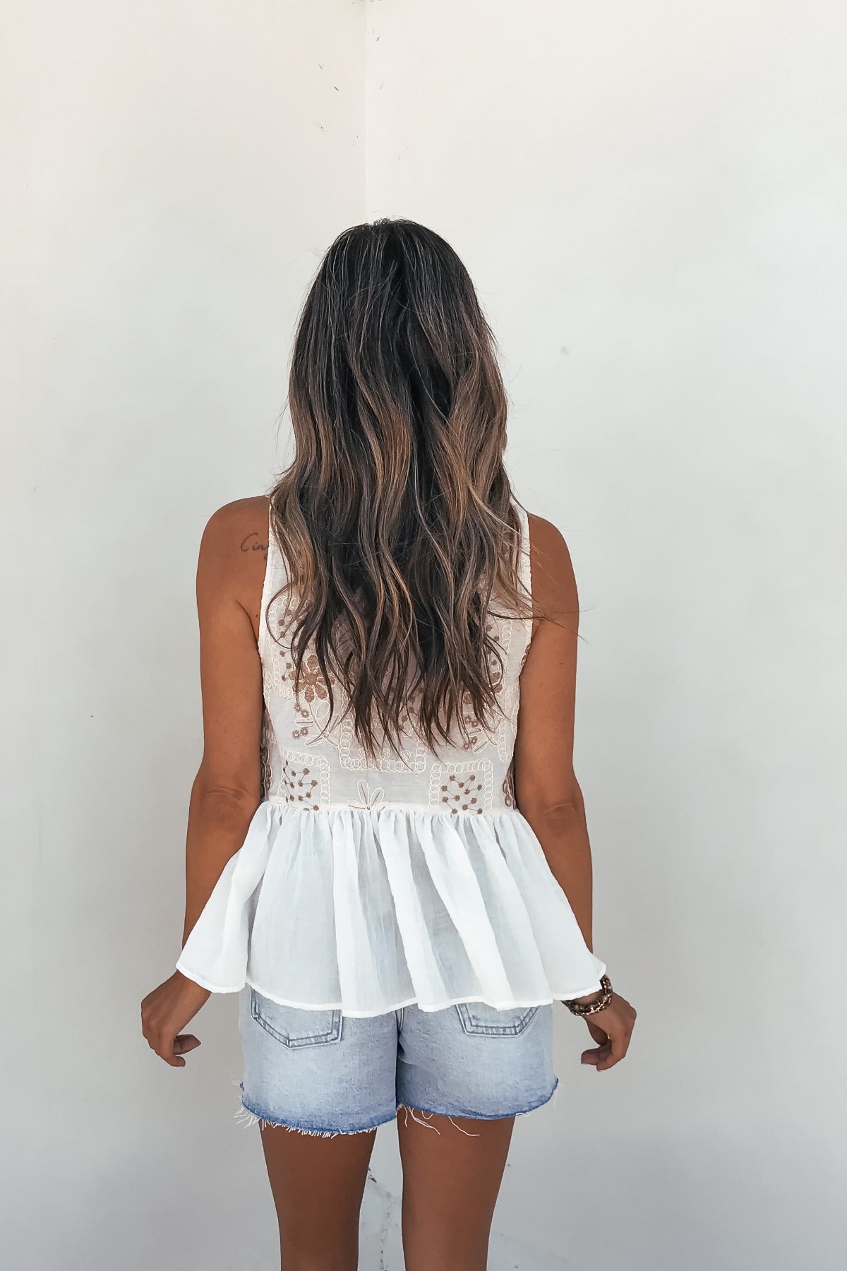 Woman with long wavy hair in the Aveline Corset Tank Top - Oatmeal, highlighting its flared hem, facing away by a white wall.