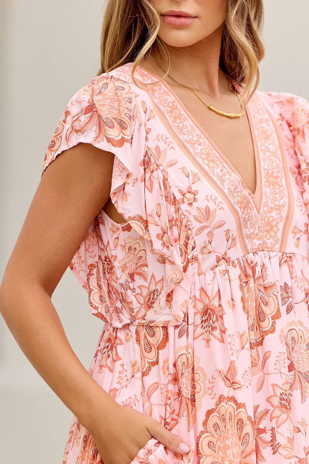 Woman in the Blush Floral Print Mini Dress-FINAL SALE, with ruffle detail, v-neckline, wavy light brown hair, and gold necklace.