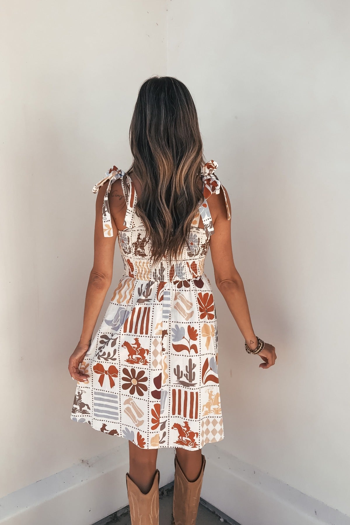 Woman with long wavy hair wears the Natural Print Mini Dress—FINAL SALE—facing a white wall, back of dress visible.