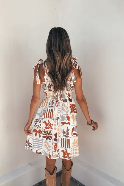 Woman with long wavy hair wears the Natural Print Mini Dress—FINAL SALE—facing a white wall, back of dress visible.