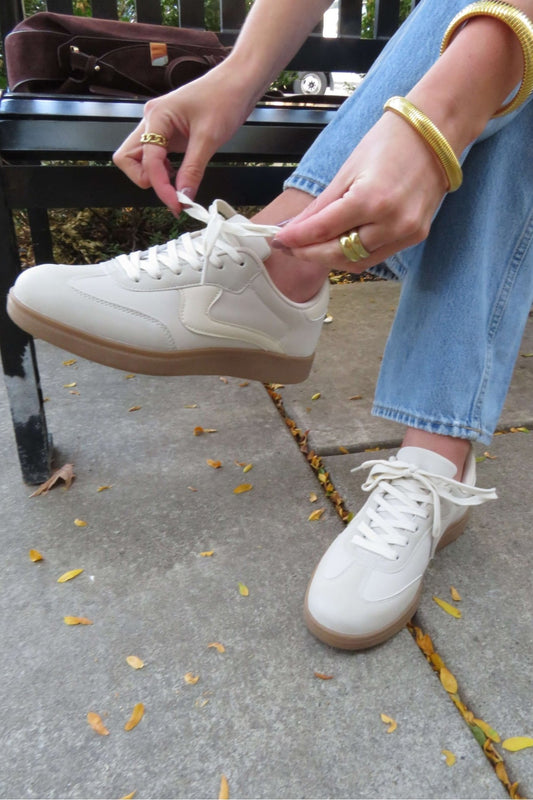 Person tying the laces of Beige Remus Lace Up Sneakers while sitting on a bench, wearing blue jeans and gold jewelry.