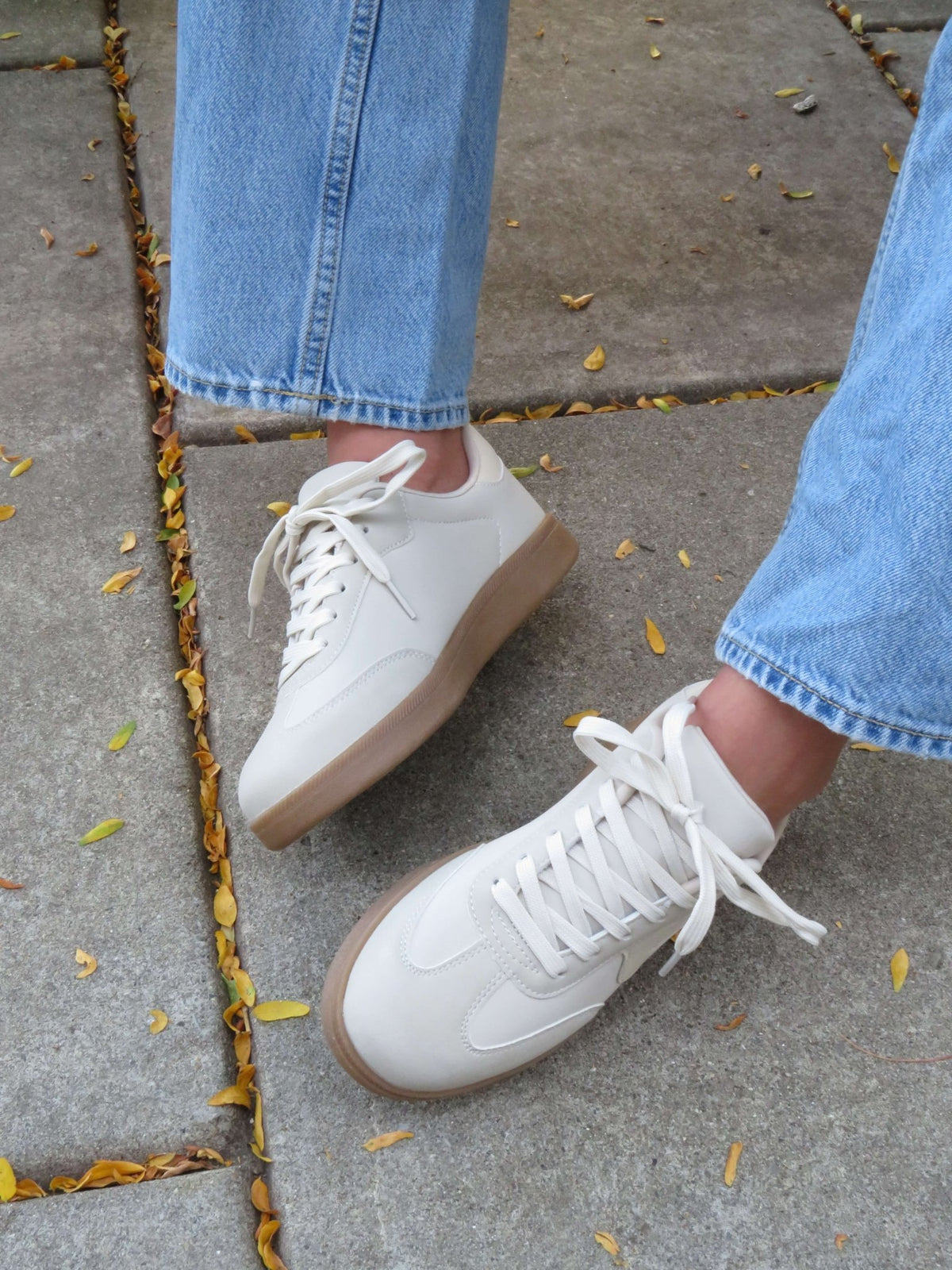 Person in light blue jeans and Beige Remus Lace Up Sneakers - FINAL SALE stands on stone pavement with yellow leaves.