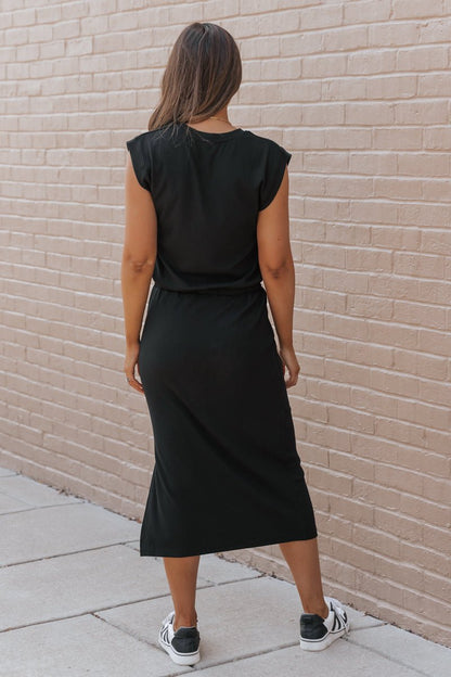 A woman in a Black Drawstring Slit Midi Dress-FINAL SALE and sneakers faces a tan brick wall, her long hair flowing behind.
