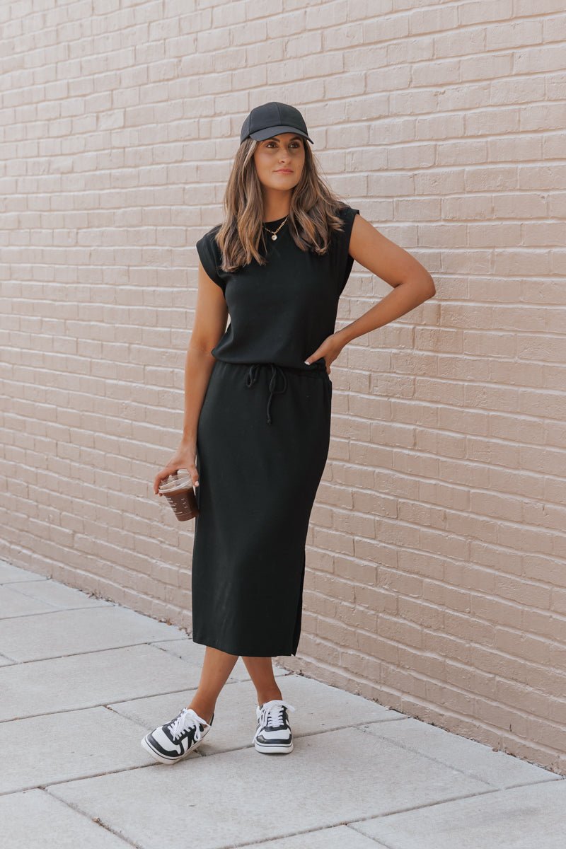 Woman in Black Drawstring Slit Midi Dress-FINAL SALE, black cap & sneakers, stands by beige brick wall holding iced coffee.
