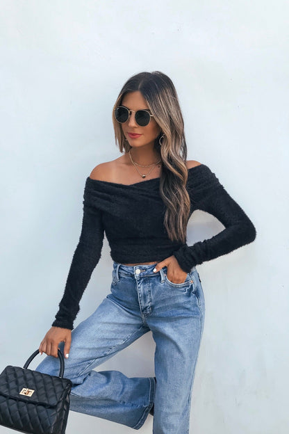 Woman in sunglasses and jeans models the Black Fuzzy Knit Off-Shoulder Top, holding a black handbag against a white backdrop.