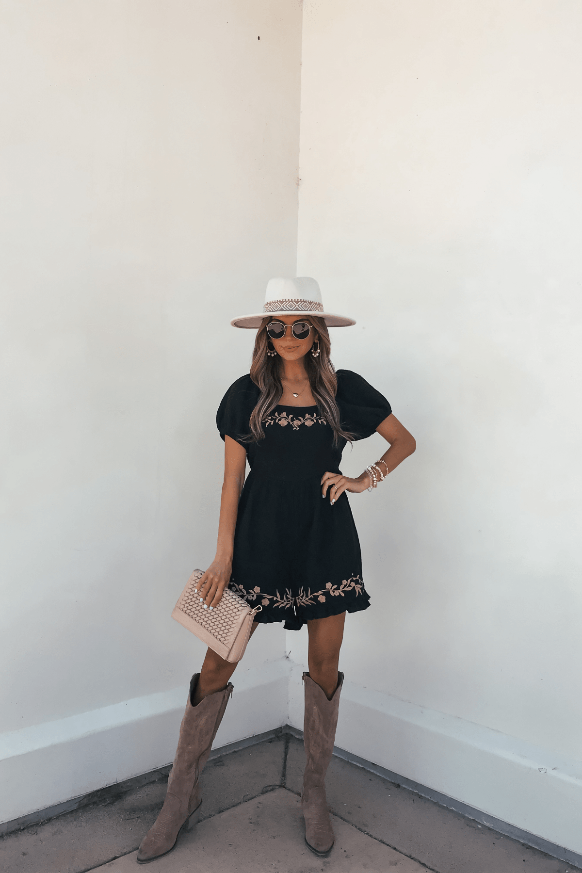 Woman in a Black Puff Sleeve Embroidered Romper, wide-brim hat, sunglasses, clutch, and boots stands against a white wall.