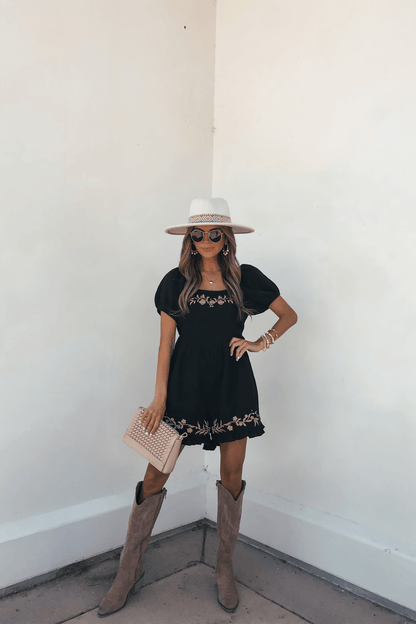 Woman in a Black Puff Sleeve Embroidered Romper, wide-brim hat, sunglasses, clutch, and boots stands against a white wall.