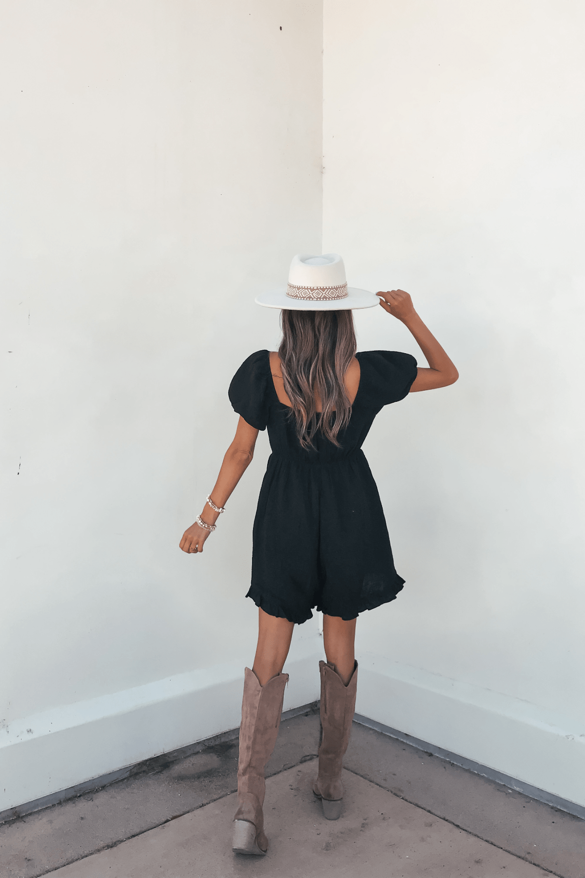 A woman in the Black Puff Sleeve Embroidered Romper-FINAL SALE and cowboy boots holds a white hat, facing a white wall.