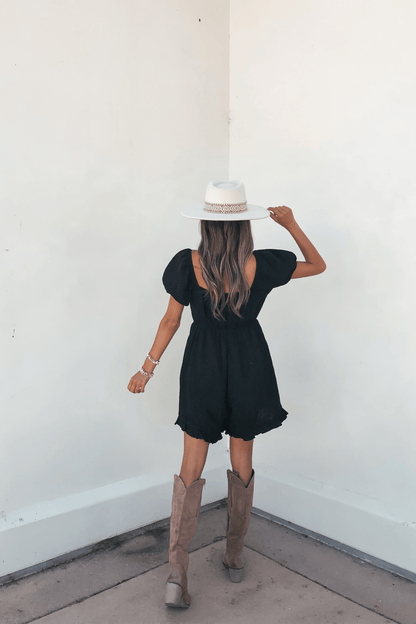 A woman in the Black Puff Sleeve Embroidered Romper-FINAL SALE and cowboy boots holds a white hat, facing a white wall.