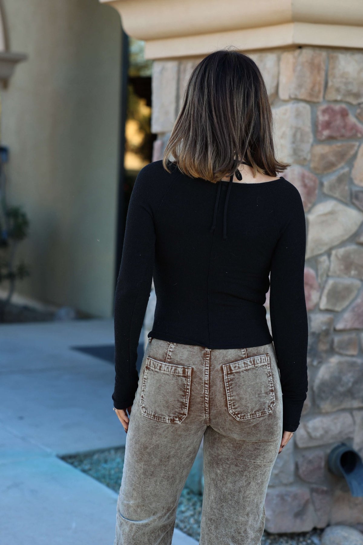 Someone wearing the Black Ruched Long Sleeve Top - FINAL SALE pairs it with brown jeans, standing outdoors facing away.
