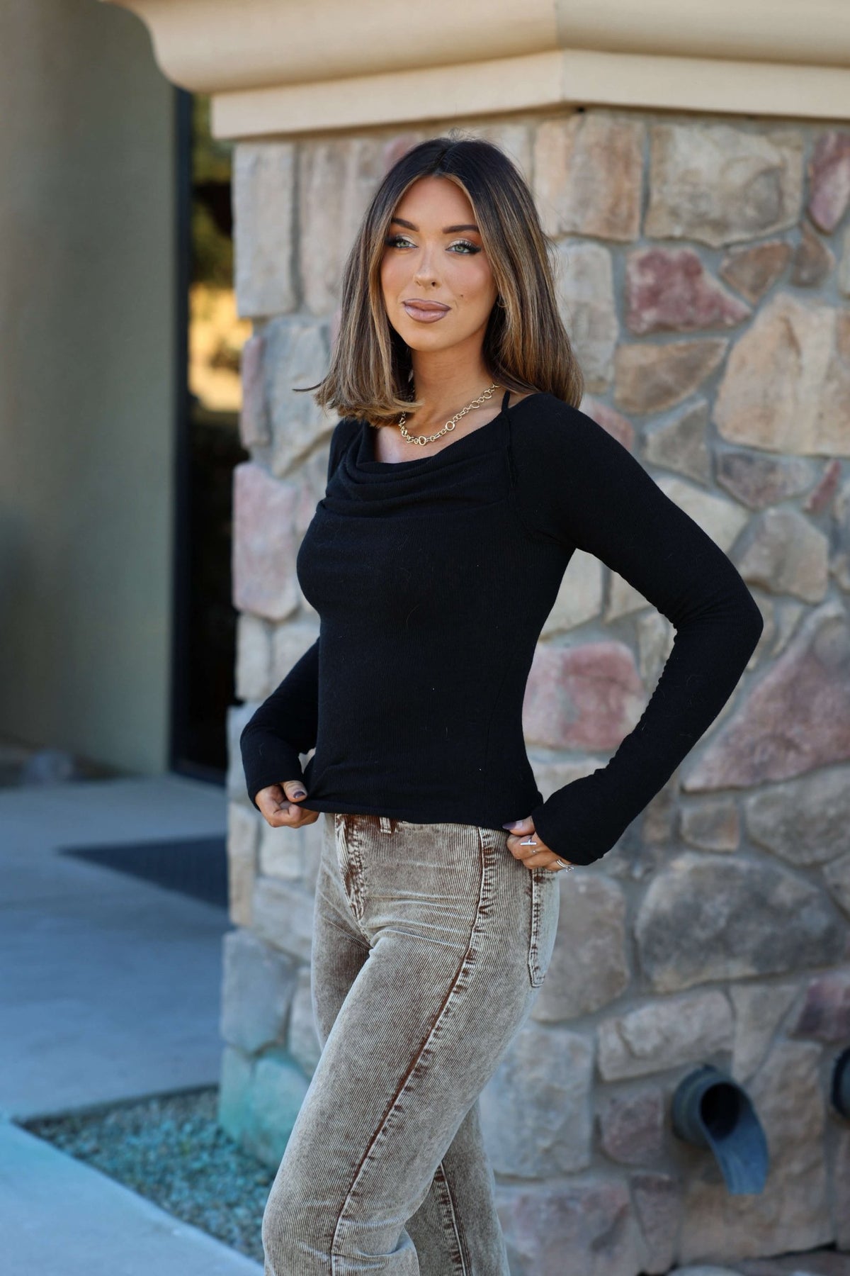 Smiling woman with shoulder-length hair wears the Black Ruched Long Sleeve Top - FINAL SALE and beige jeans by a stone wall.
