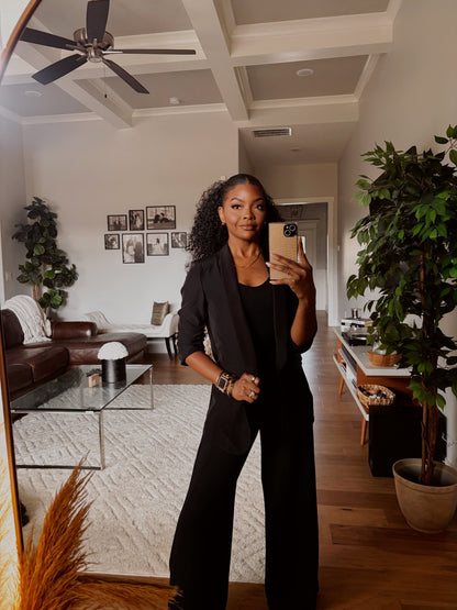 A woman in a Black Ruched Sleeve Blazer takes a mirror selfie in a modern living room with plants and wall art in the background.
