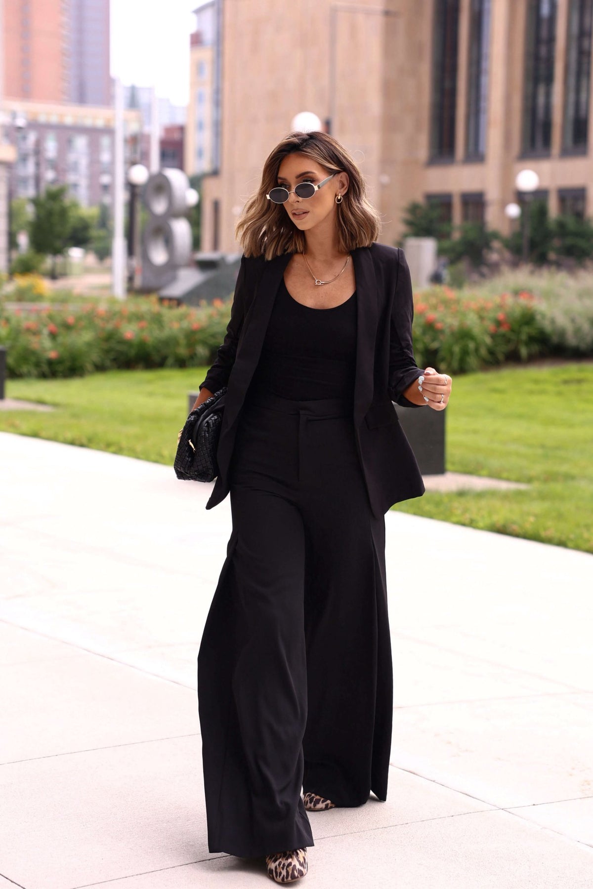 A woman in a Black Ruched Sleeve Blazer walks outside on a city sidewalk with greenery and buildings in the background.