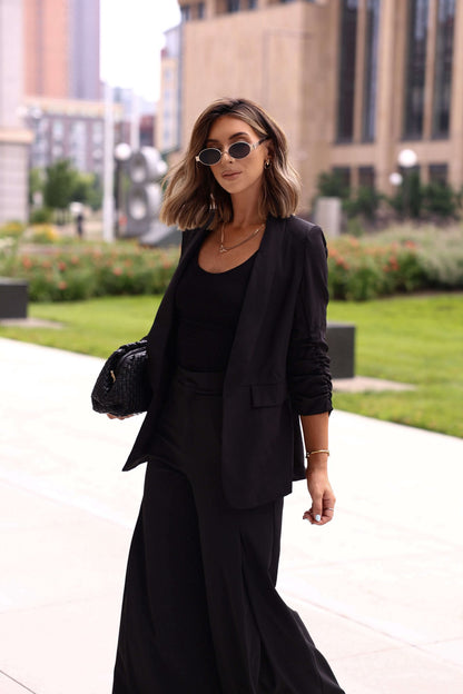 Office-ready: Woman in a Black Ruched Sleeve Blazer, black top, wide-leg pants, sunglasses, and black handbag, walking outdoors.