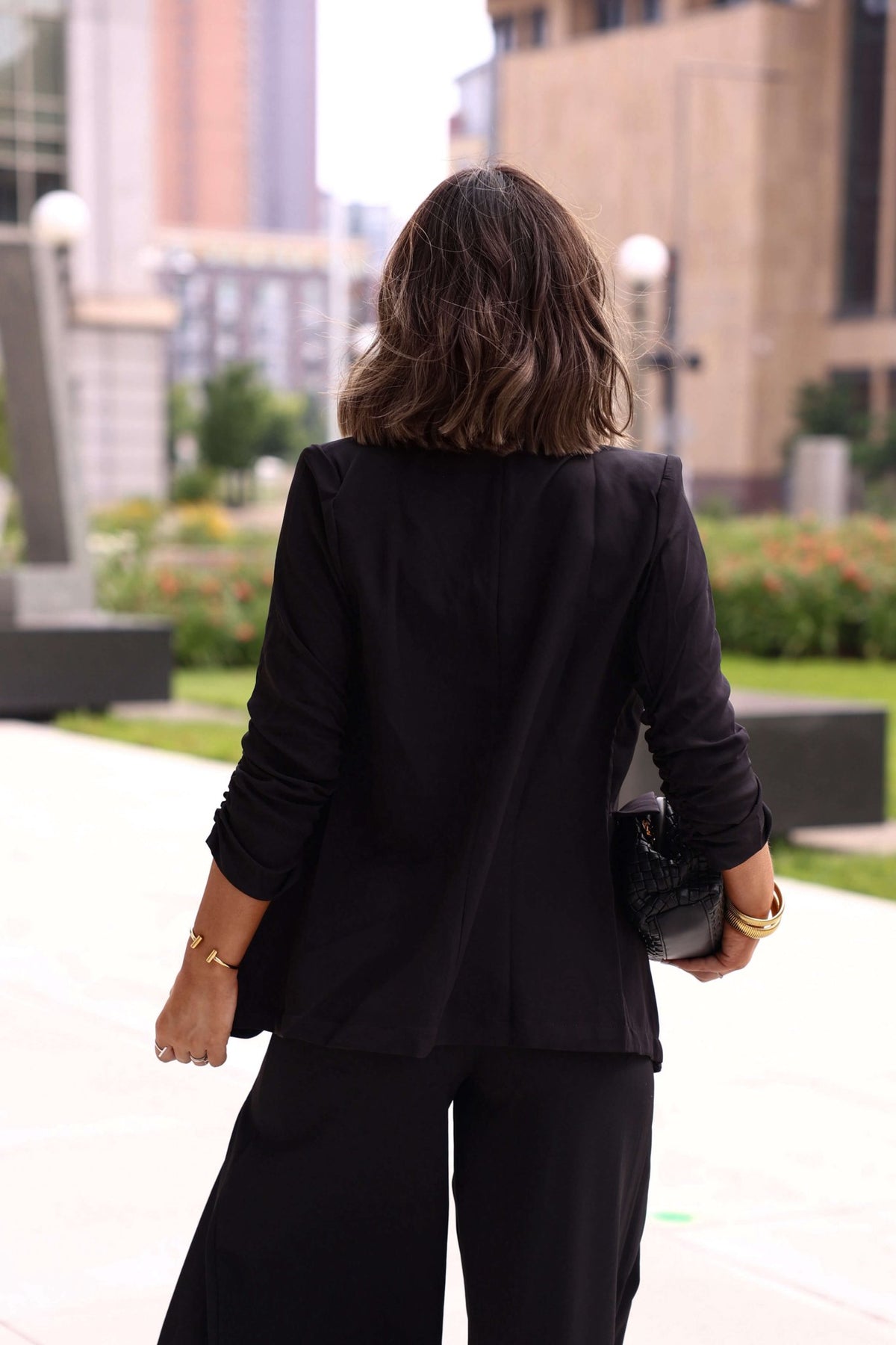 A woman with shoulder-length hair wears the Black Ruched Sleeve Blazer outdoors, holding a black clutch for an office-ready look.