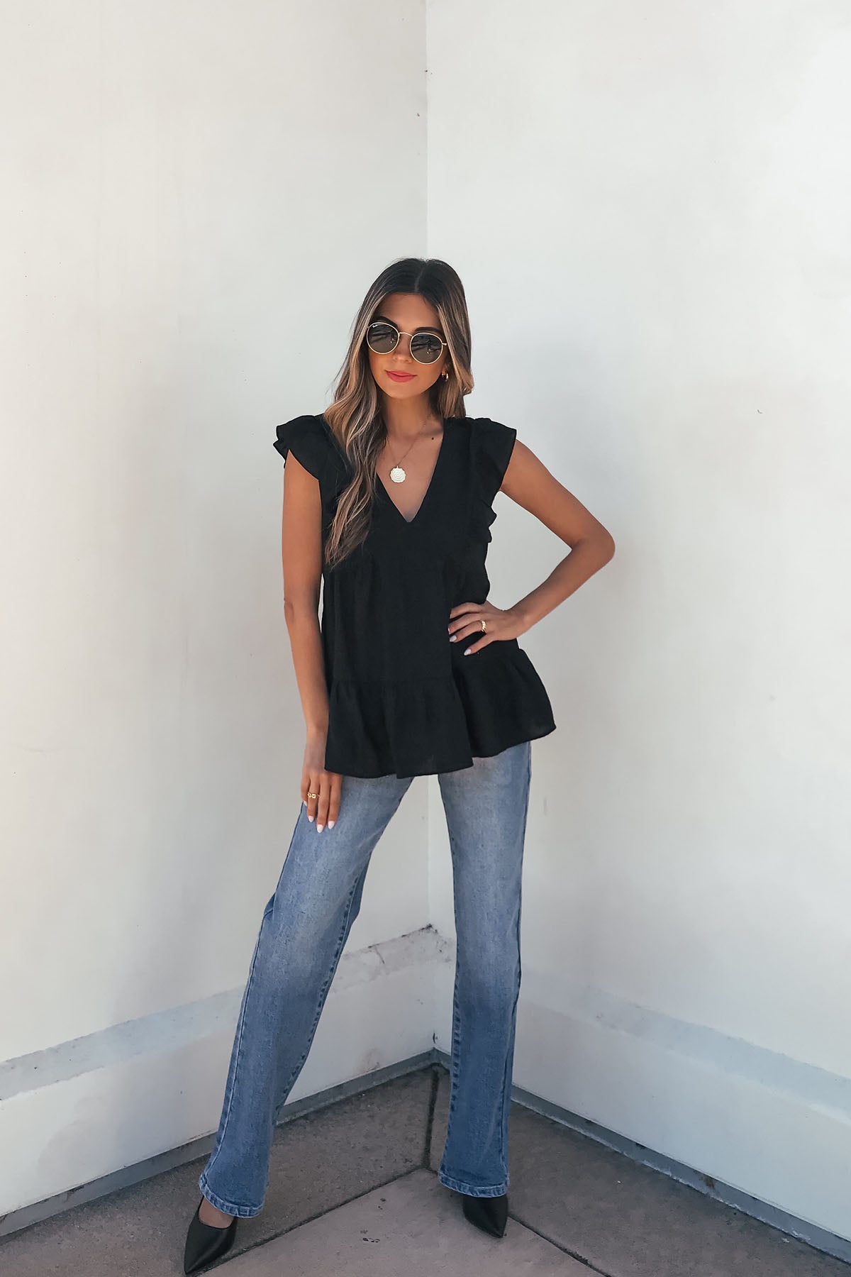 Woman wearing the Black Ruffle Sleeve Textured Top- FINAL SALE, blue jeans, and black heels stands confidently by a white wall.