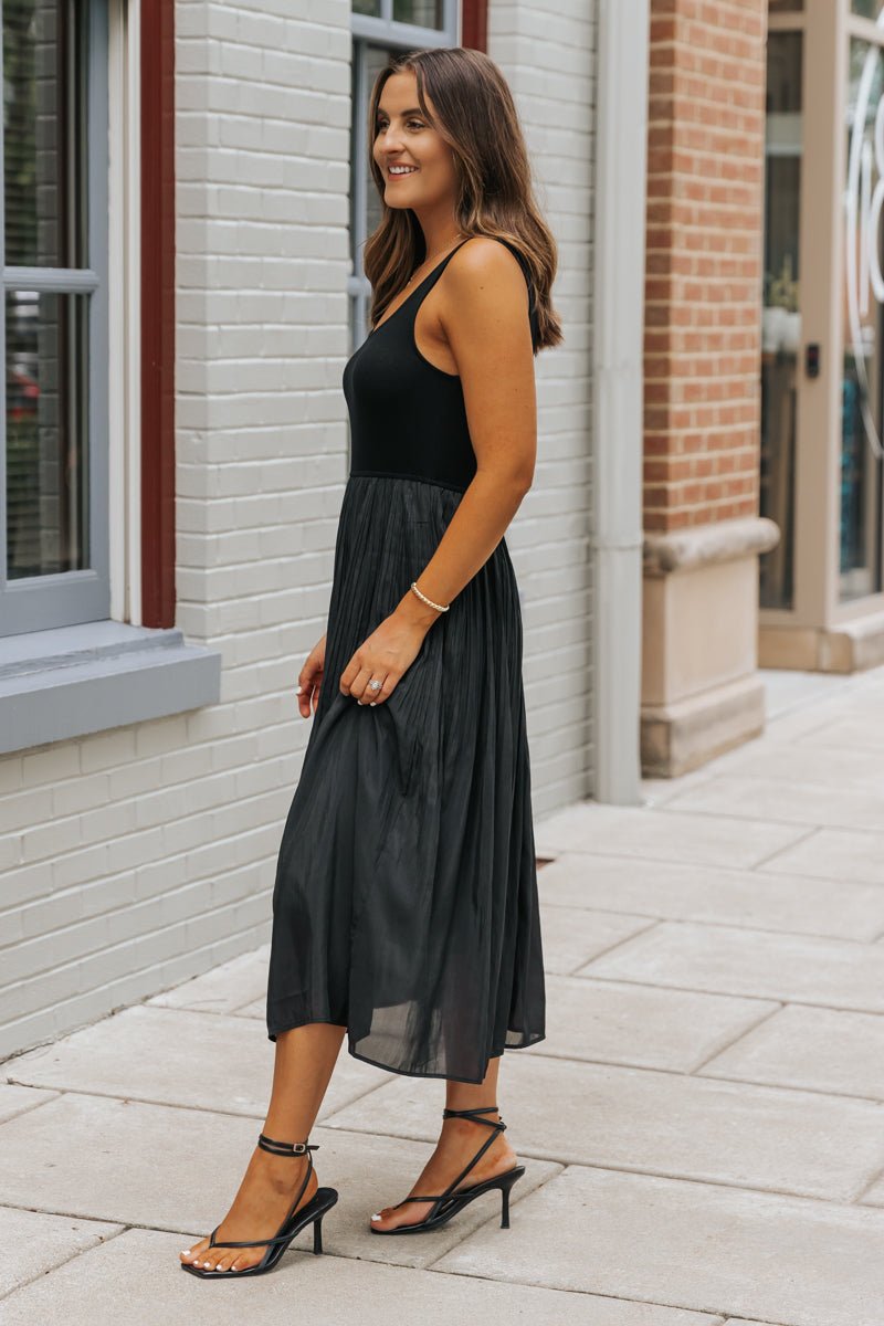 Woman in the Black Scoop Neck Pleated Tank Midi Dress stands on a sidewalk by brick buildings, smiling.