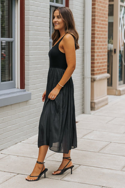 Woman in the Black Scoop Neck Pleated Tank Midi Dress stands on a sidewalk by brick buildings, smiling.