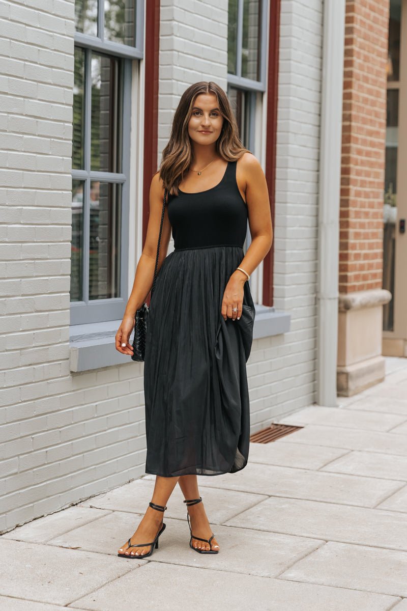 Woman in the Black Scoop Neck Pleated Tank Midi Dress stands on a city sidewalk, styled with black heels and a chic bag.