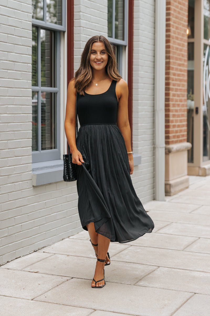 Woman wearing the Black Scoop Neck Pleated Tank Midi Dress, smiling as she walks in sandals by a brick building.