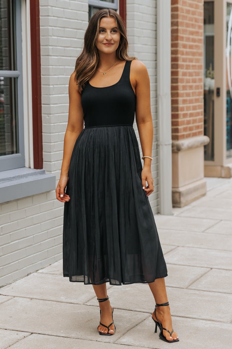 Woman wearing a Black Scoop Neck Pleated Tank Midi Dress stands on a city sidewalk, smiling at the camera.