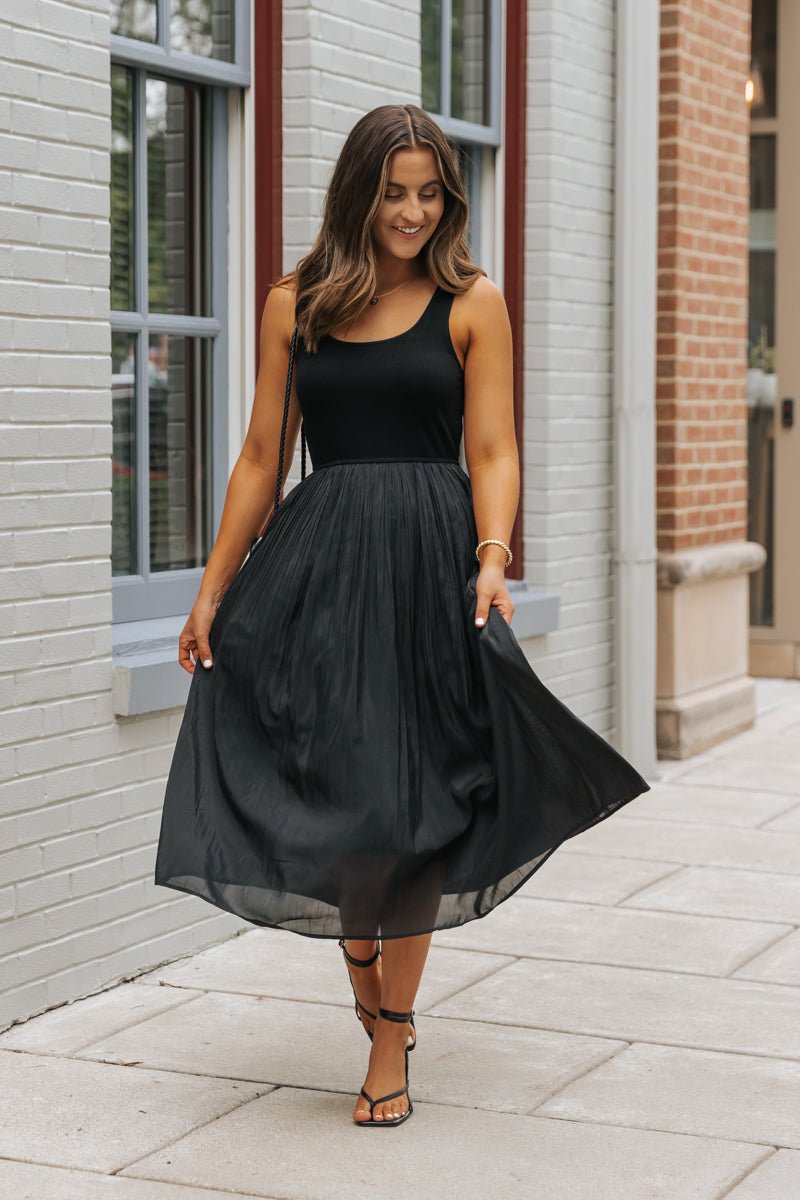 Woman in the Black Scoop Neck Pleated Tank Midi Dress, smiling and walking, holding her midi skirt.