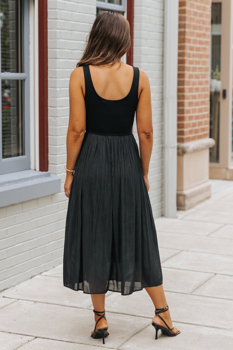 Woman wears the Black Scoop Neck Pleated Tank Midi Dress-FINAL SALE with heels, standing on a sidewalk by brick and painted buildings.