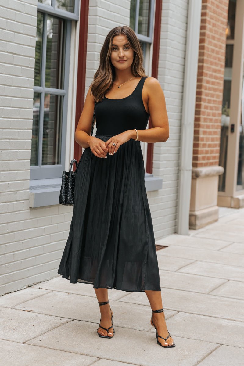 Woman in a Black Scoop Neck Pleated Tank Midi Dress stands on city sidewalk, holding a black handbag and wearing sandals.