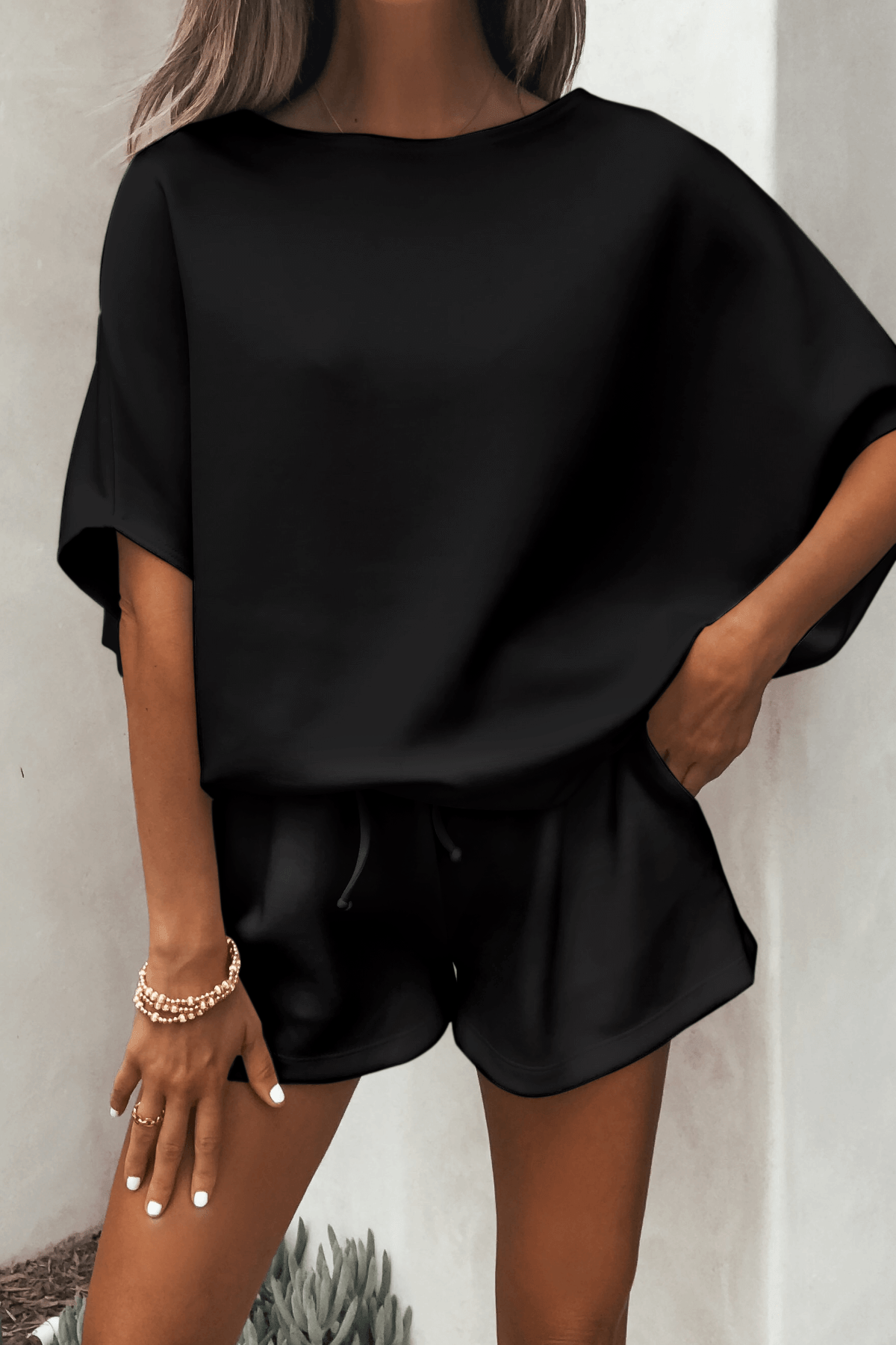 Woman in a Black Soft Cloud Scuba Top and black shorts, gold bracelets on her wrist, stands against a light wall.
