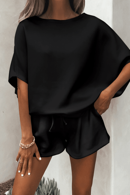 Woman in a Black Soft Cloud Scuba Top and black shorts, gold bracelets on her wrist, stands against a light wall.