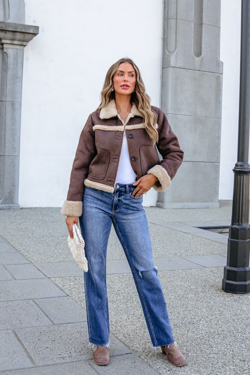Woman in a BLU PEPPER TW2302 BROWN JACKET, white top, jeans, and boots stands outside with a white clutch near a stone building.