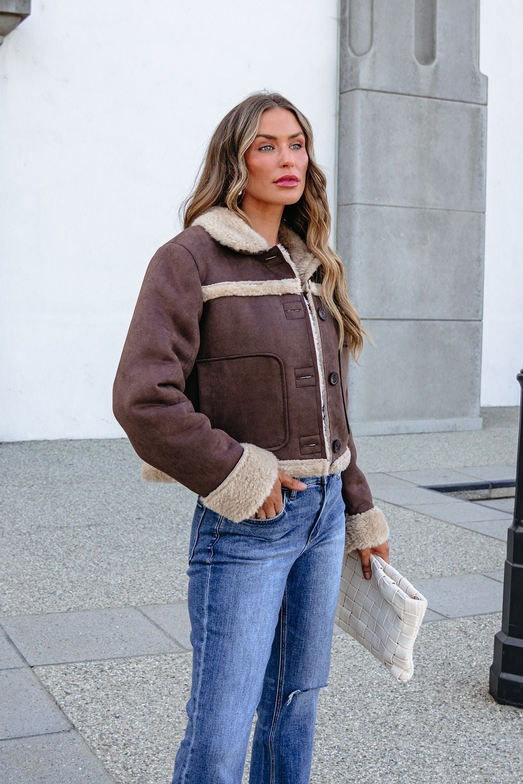 Woman in BLU PEPPER TW2302 brown jacket and jeans holds a white clutch outdoors by a gray stone wall.
