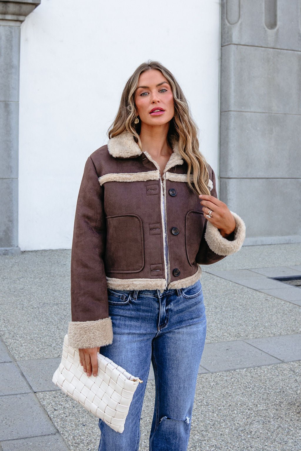 Woman in a BLU PEPPER TW2302 brown jacket, blue jeans, holding a white clutch, stands outside by a stone wall.