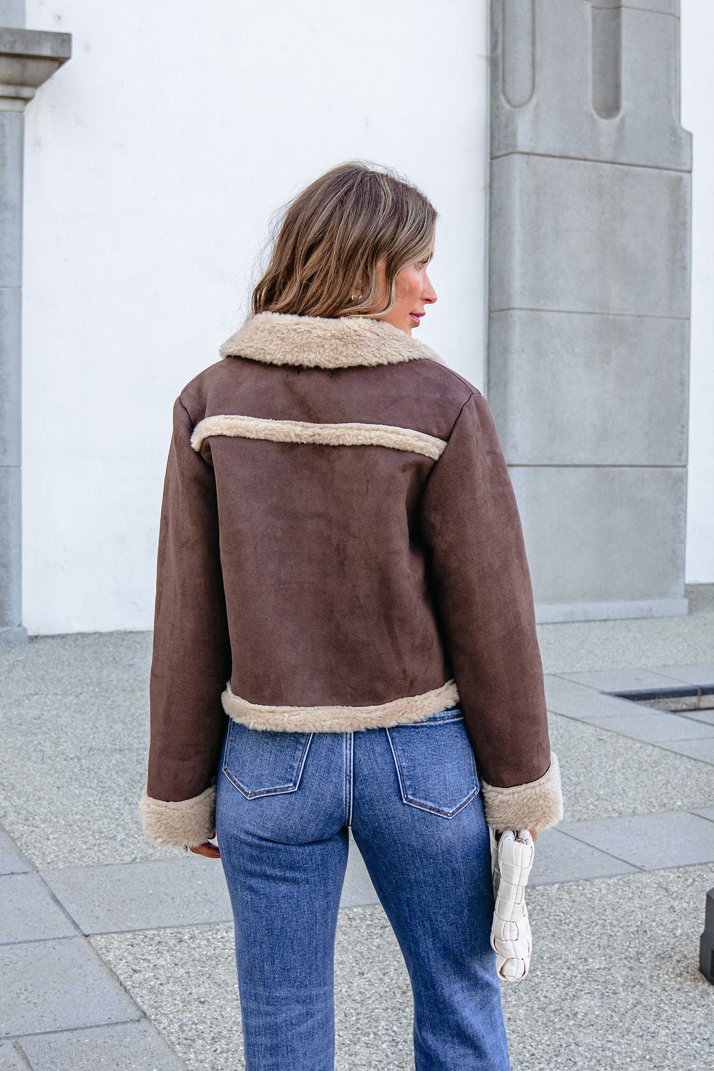 A woman in the BLU PEPPER TW2302 Brown Jacket and blue jeans stands outdoors, facing away, holding a small white purse.