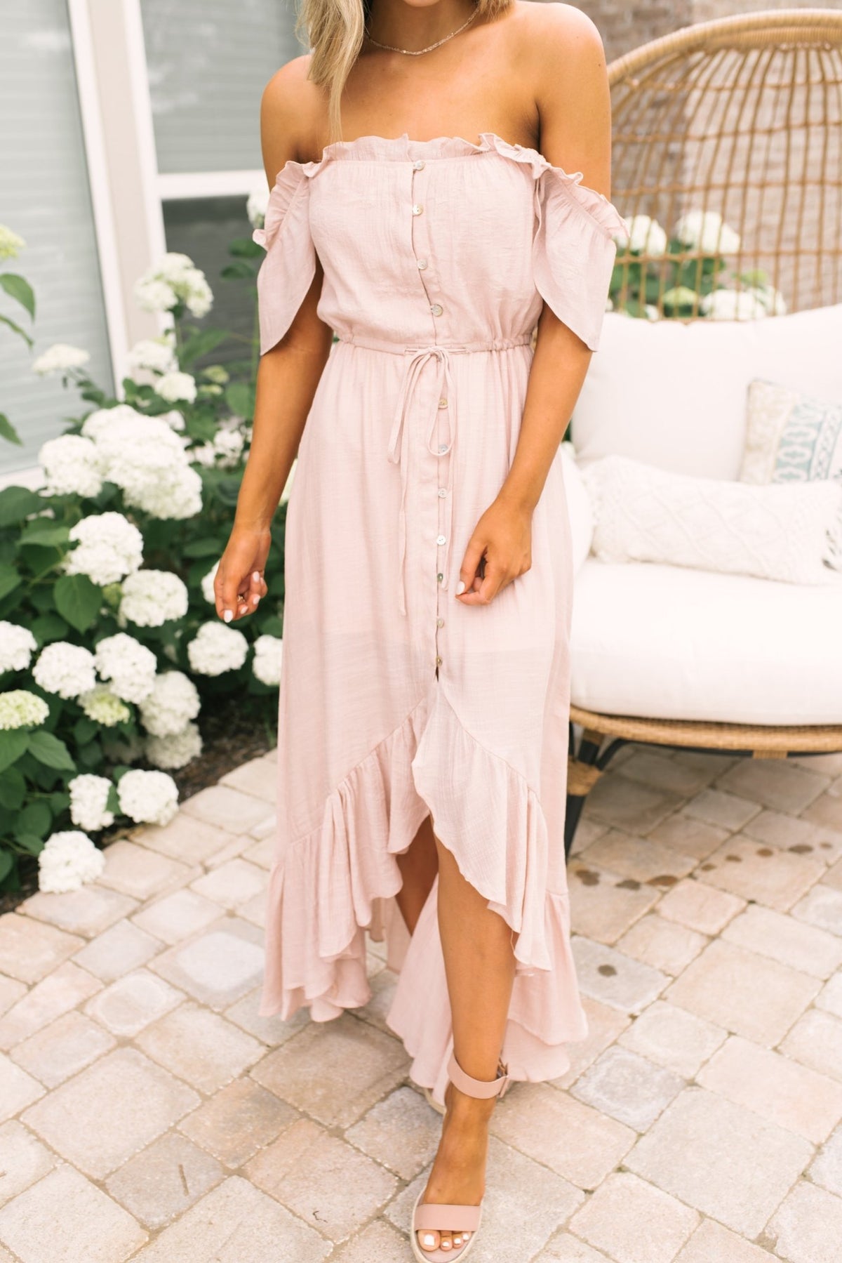 Woman in Blush Off the Shoulder Button Down High Low Maxi stands on a patio near white flowers and a cushioned chair.