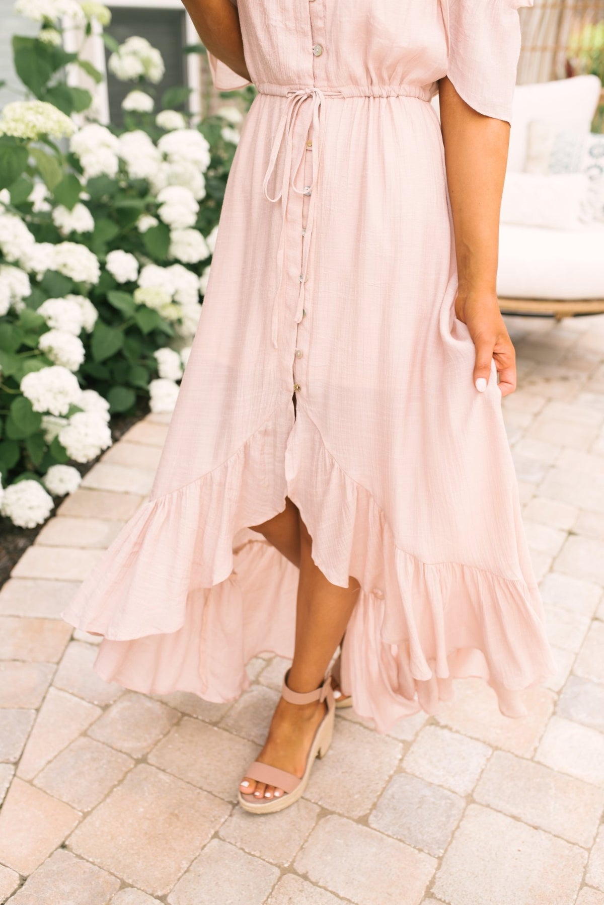Woman in a Blush Off the Shoulder Button Down High Low Maxi stands on a stone patio near blooming white flowers.