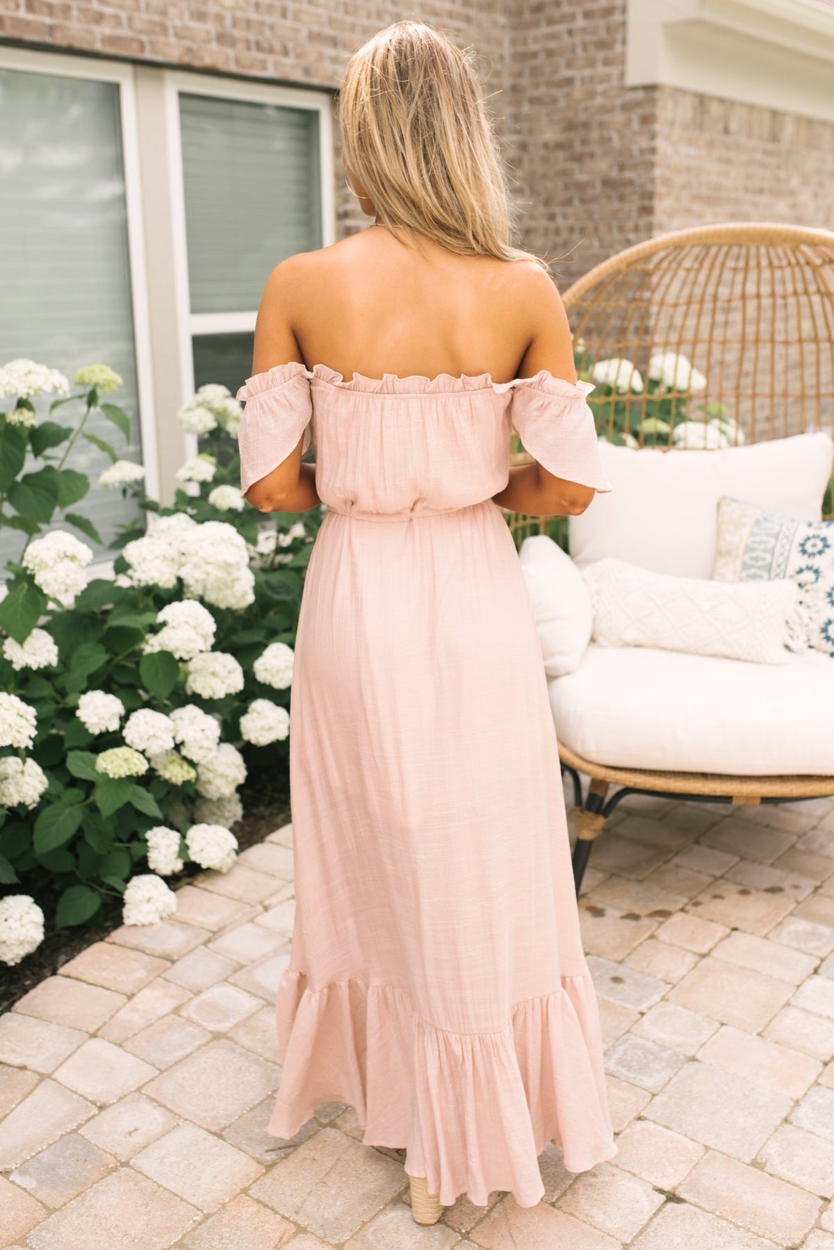 Woman in Blush Off the Shoulder Button Down High Low Maxi stands on a patio near white flowers; ideal for wedding guests.