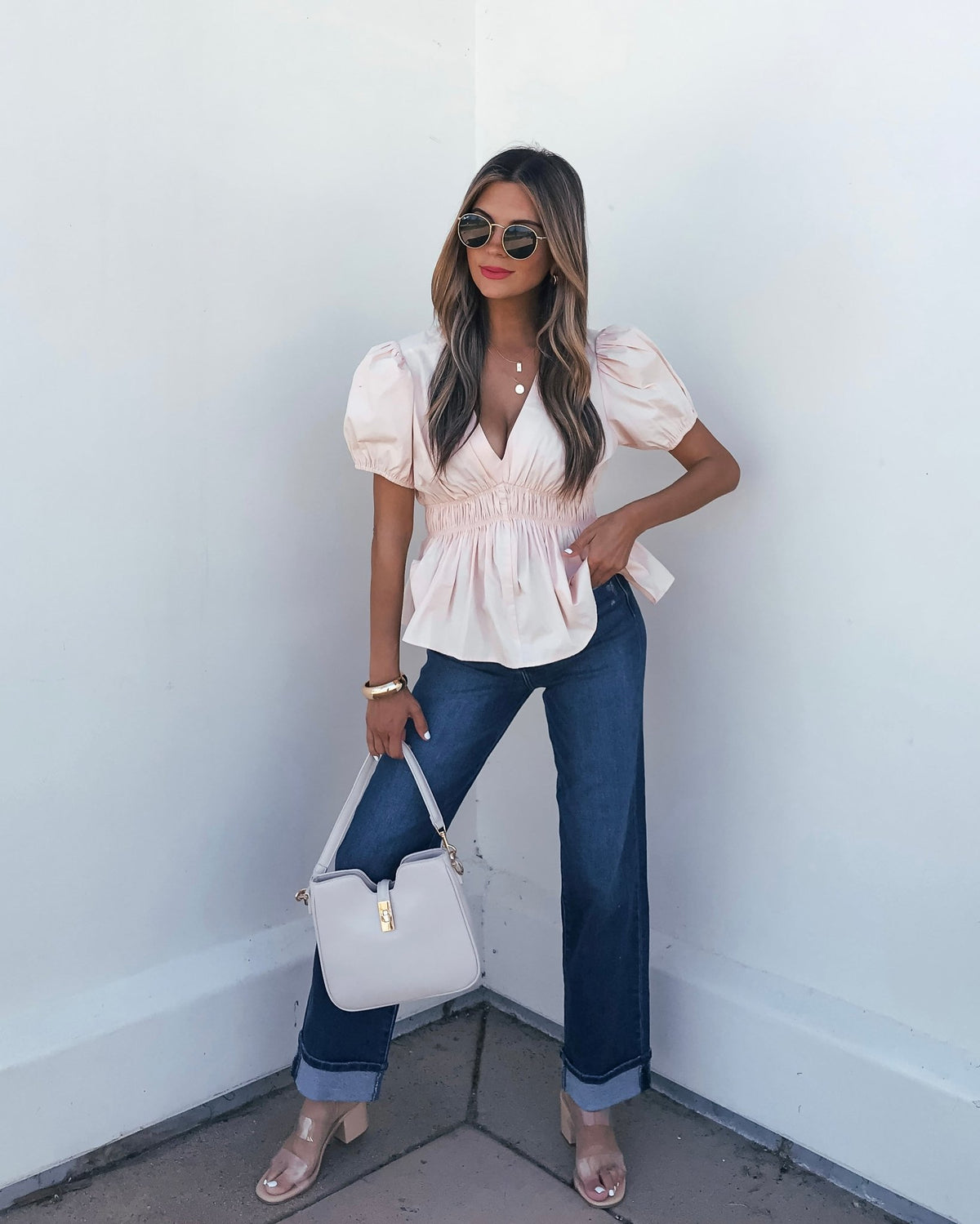 Woman in sunglasses wears the Blush Puff Sleeve Peplum Top, blue jeans, nude heels, and holds a beige purse against a white wall.