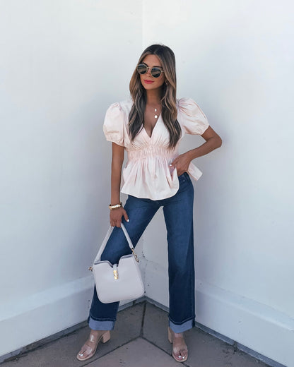 Woman in sunglasses wears the Blush Puff Sleeve Peplum Top, blue jeans, nude heels, and holds a beige purse against a white wall.
