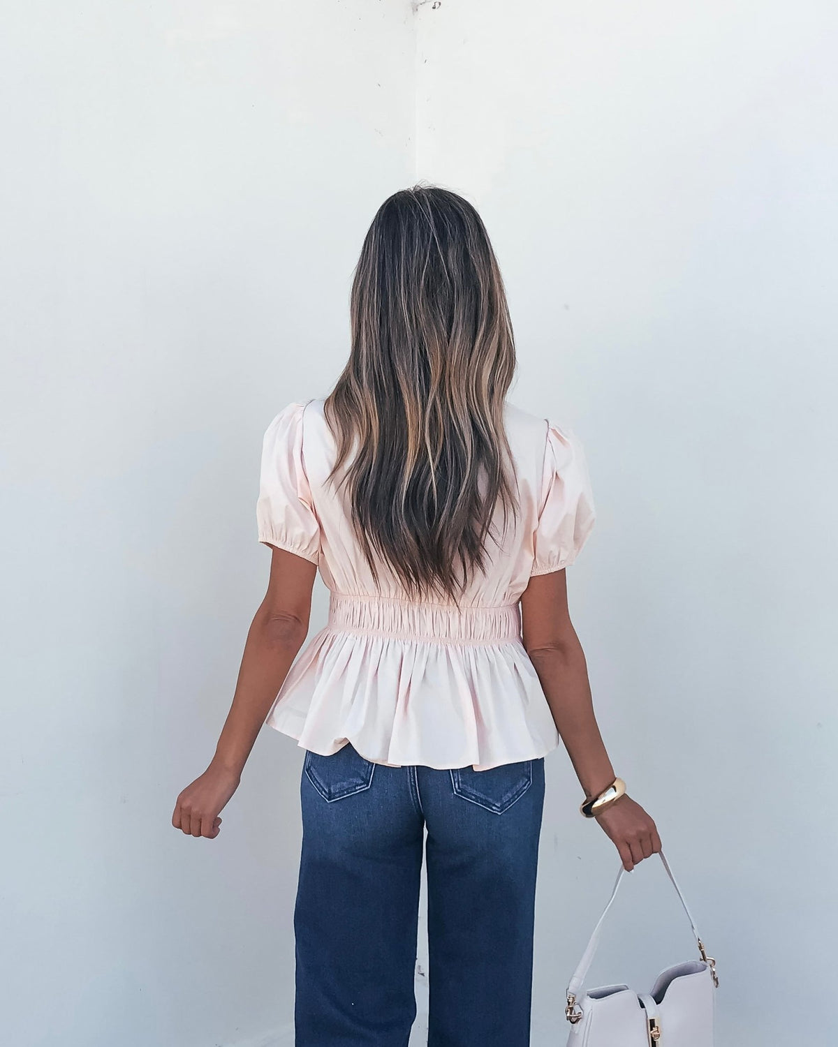 Woman with long wavy hair wears the Blush Puff Sleeve Peplum Top and jeans, facing a white wall, holding a pale handbag in her right hand.