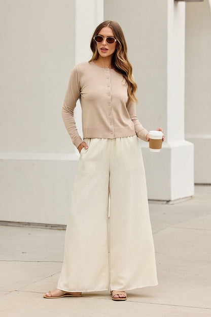 Woman in Boardroom Button Down Sweater - Tan stands outdoors with coffee, sunglasses, cream pants, and sandals.