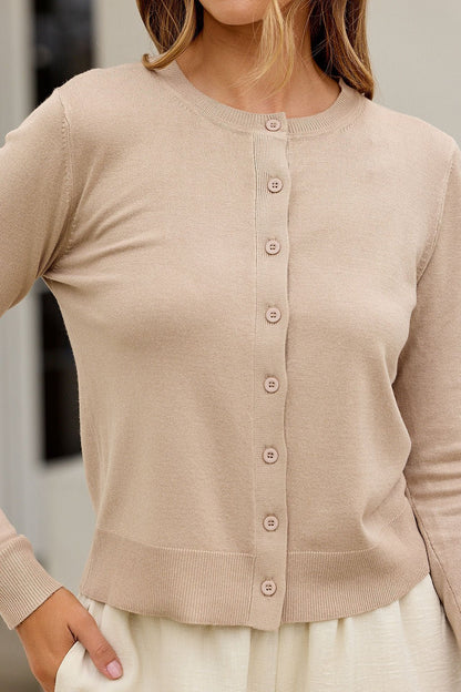 Woman in a tan Boardroom Button Down Sweater - FINAL SALE and cream skirt, standing with one hand on her hip.
