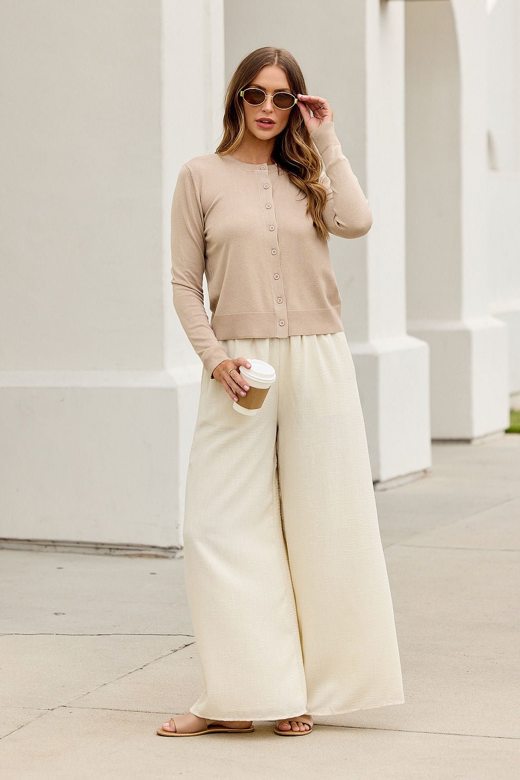Woman in Boardroom Button Down Sweater - Tan holds coffee, standing on a sidewalk next to a white building.