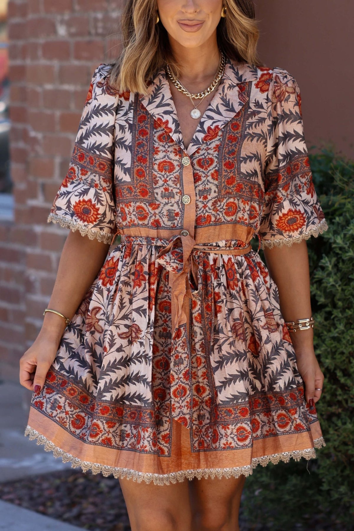 Woman wearing the Botanical Rust Print Belted Mini Dress stands by a brick wall and greenery—perfect for your fall wardrobe.