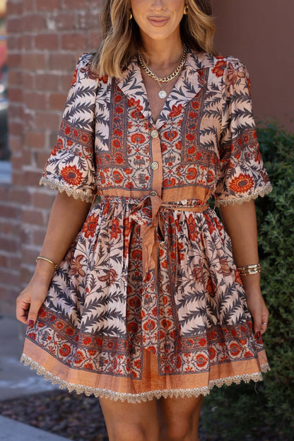 Woman wearing the Botanical Rust Print Belted Mini Dress stands by a brick wall and greenery—perfect for your fall wardrobe.