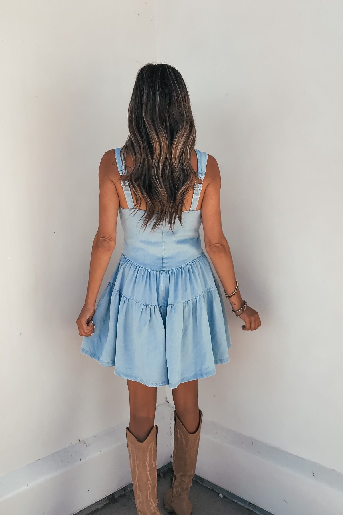 Woman with wavy hair in Briar Chambray Tiered Romper-FINAL SALE and tan cowboy boots, back facing the camera.