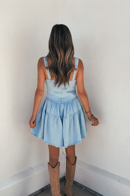 Woman with wavy hair in Briar Chambray Tiered Romper-FINAL SALE and tan cowboy boots, back facing the camera.