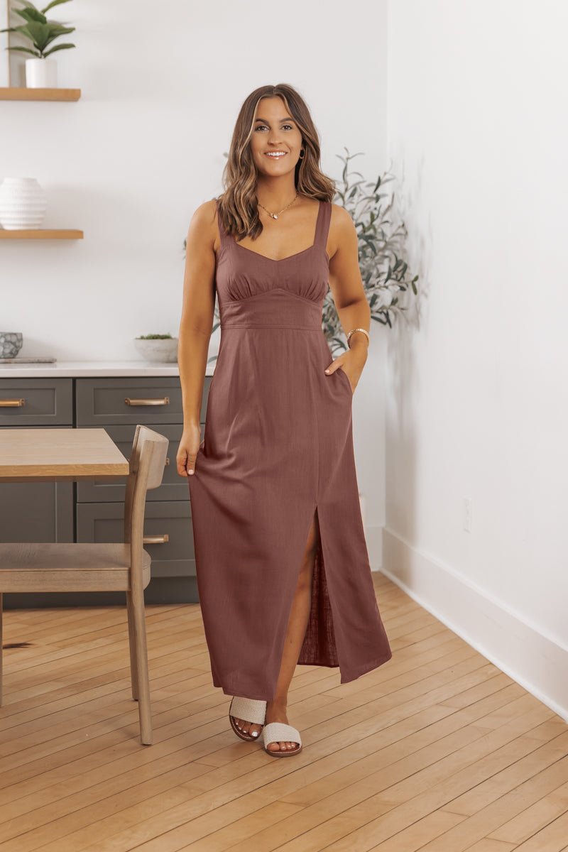 Woman wears Deep Mauve V Neck Linen Midi Dress-FINAL SALE, smiling indoors on a wood floor with one hand in her pocket.
